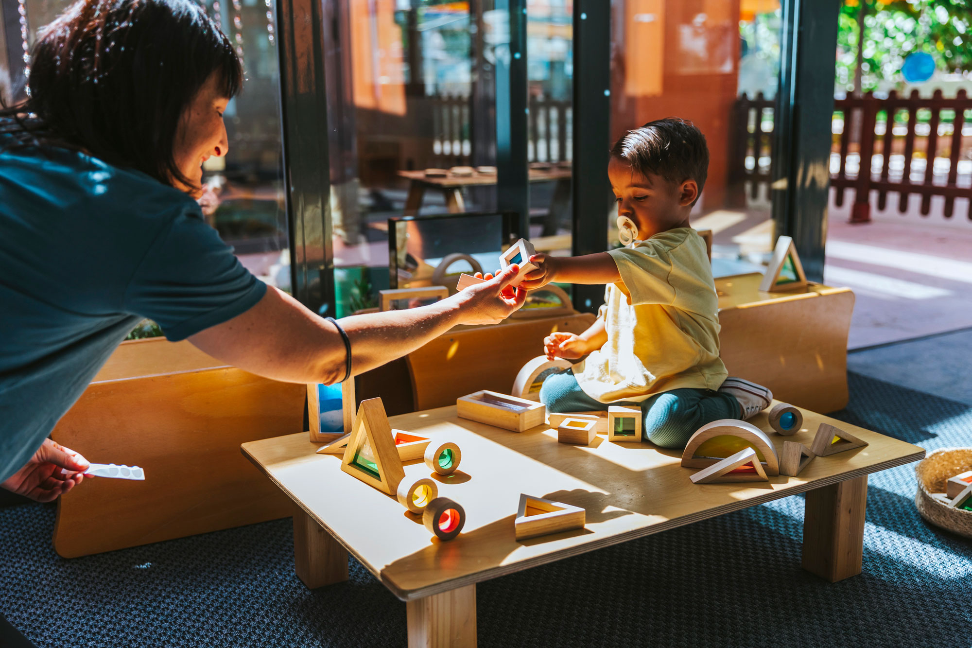 Una educadora acompaña a un niño durante el juego en un aula de educación infantil. La educadora está sentada en el suelo junto al niño, que está jugando con bloques de construcción de colores. El aula es luminosa y está equipada con juguetes educativos, estanterías de libros y muebles a medida para niños. La escena es cálida y acogedora, enfatizando un entorno educativo y de cuidado.