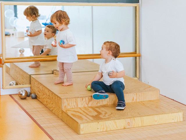 Dos niños jugando frente a un espejo en EMPI, disfrutando de una experiencia de juego sensorial y explorando su entorno en una plataforma de madera elevada.
